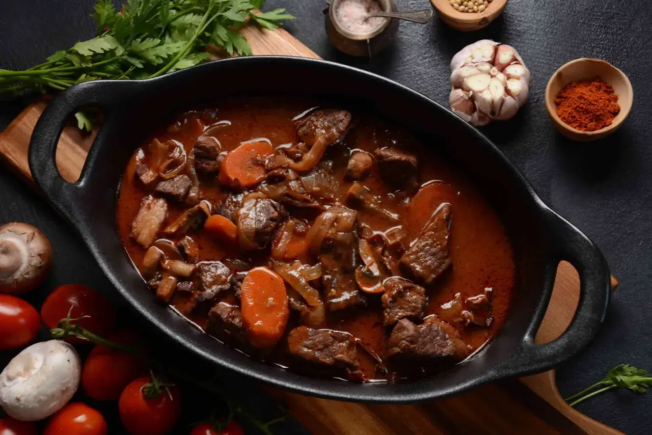 Un plat mijoté de bœuf en sauce riche, avec des carottes et des oignons. Les ingrédients pour une marinade boeuf parfaite sont disposés autour.