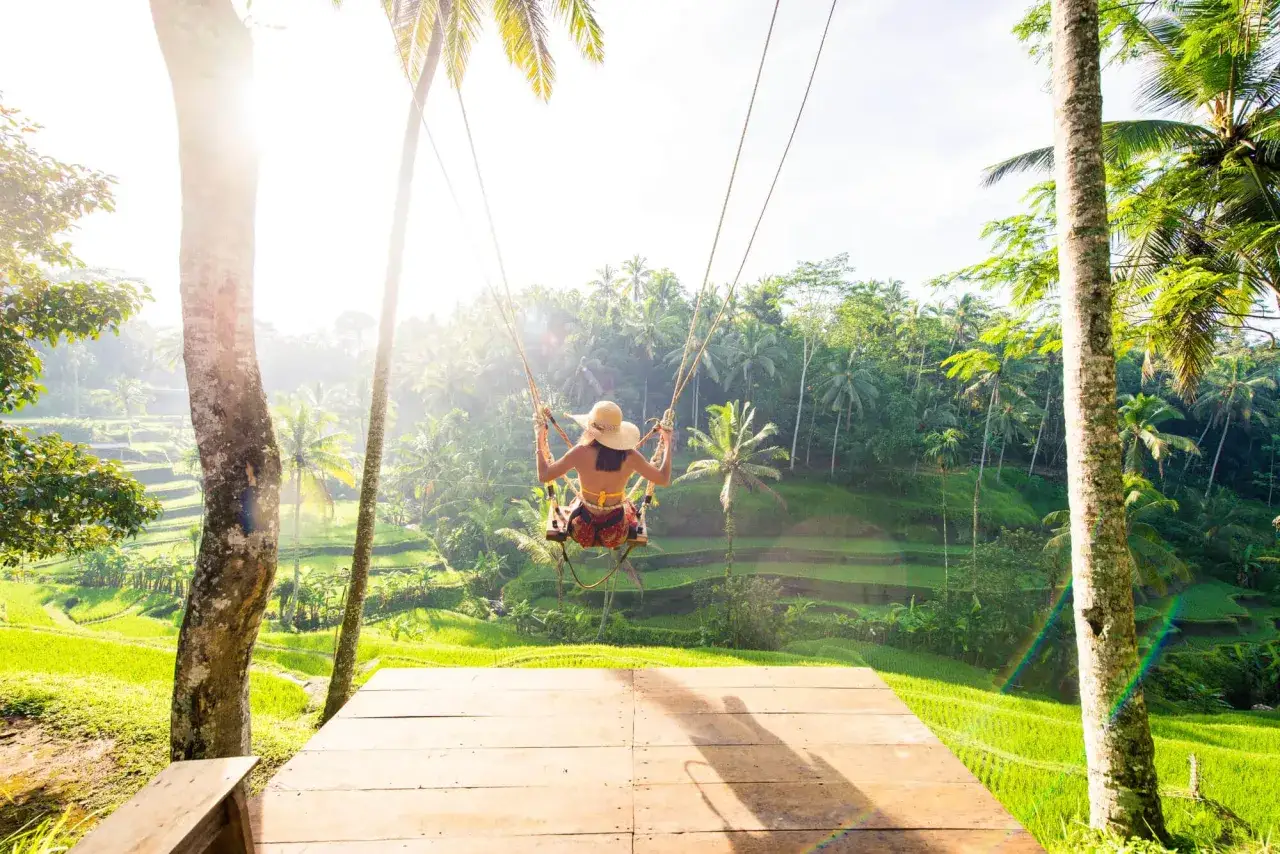 Dziewczyna na huśtawce na Bali, z widokiem na tarasowe pola ryżowe i palmy. Słońce przebija się przez liście, tworząc magiczną atmosferę.