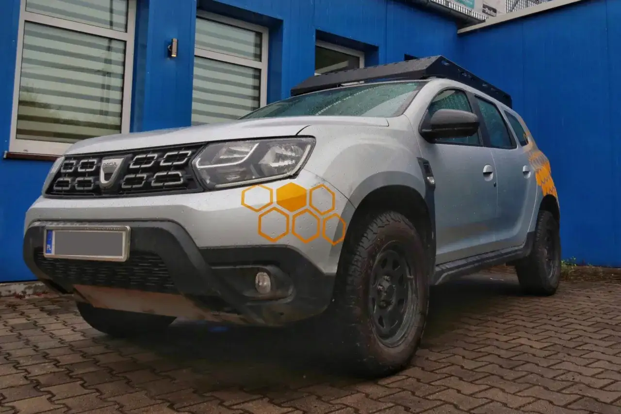 Srebrny Dacia Duster z bagażnikiem dachowym i naklejkami w kształcie plastra miodu. Idealne jakie auto na ryby, gotowe na każdą wyprawę.
