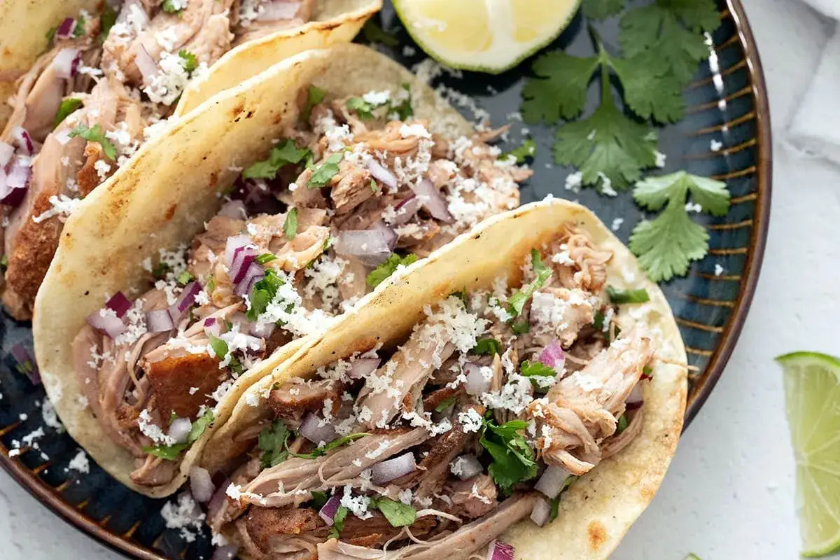 Tacos z soczystymi carnitas, posypane serem i cebulką, podane z limonką i kolendrą. Pyszne danie, które rozpływa się w ustach.