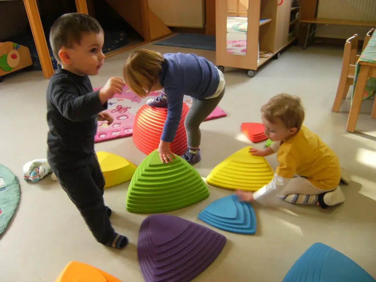 Kinder erkunden bunte Balance-Steine. Ein Junge in Schwarz balanciert, ein M&auml;dchen in Blau steht auf einem roten Podest. Offene Beobachtung im Spiel.