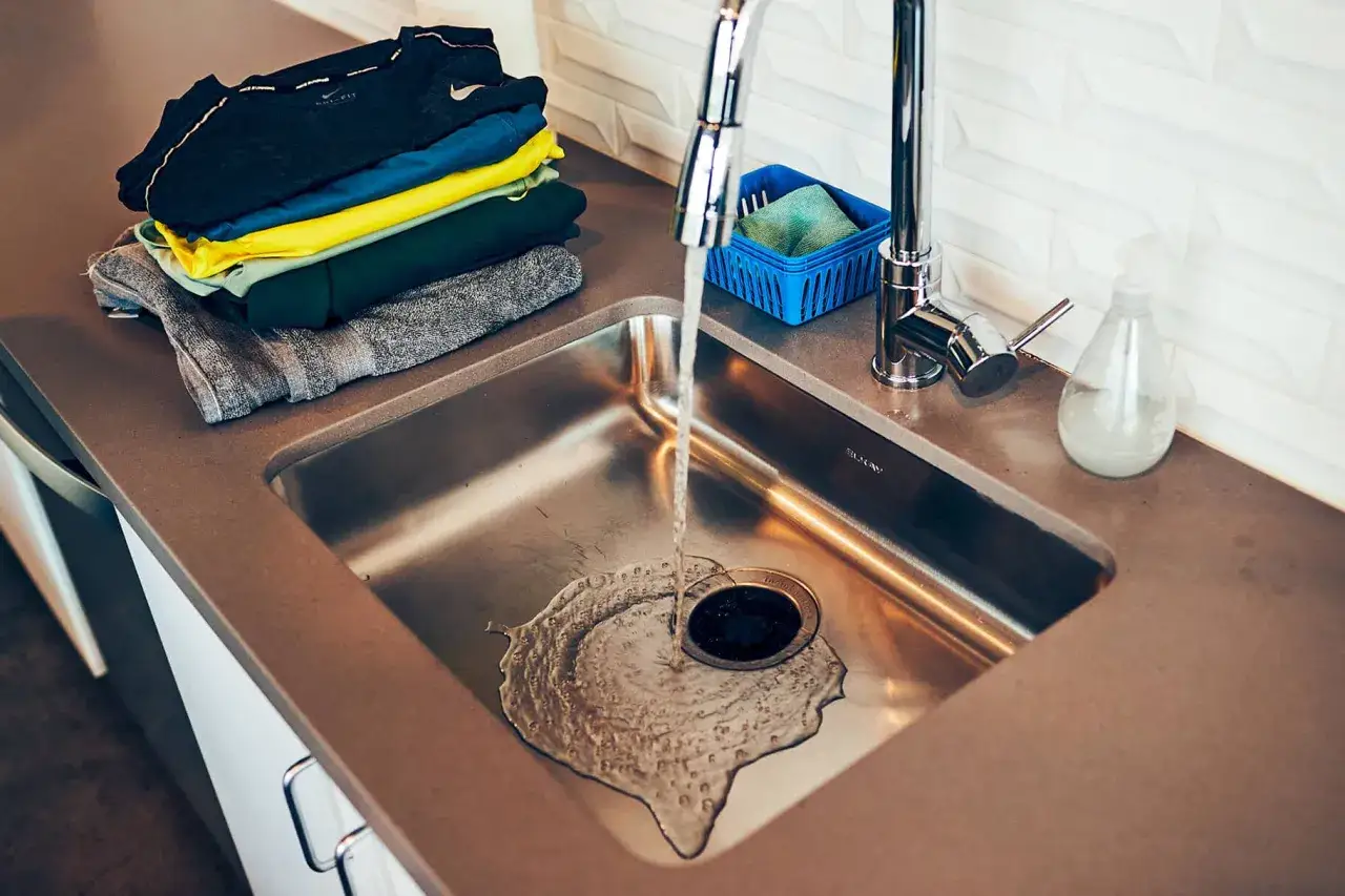 Stapel bunter Kleidung neben einer Spüle, in der Wasser läuft. So kann man auch waschen ohne waschmaschine.