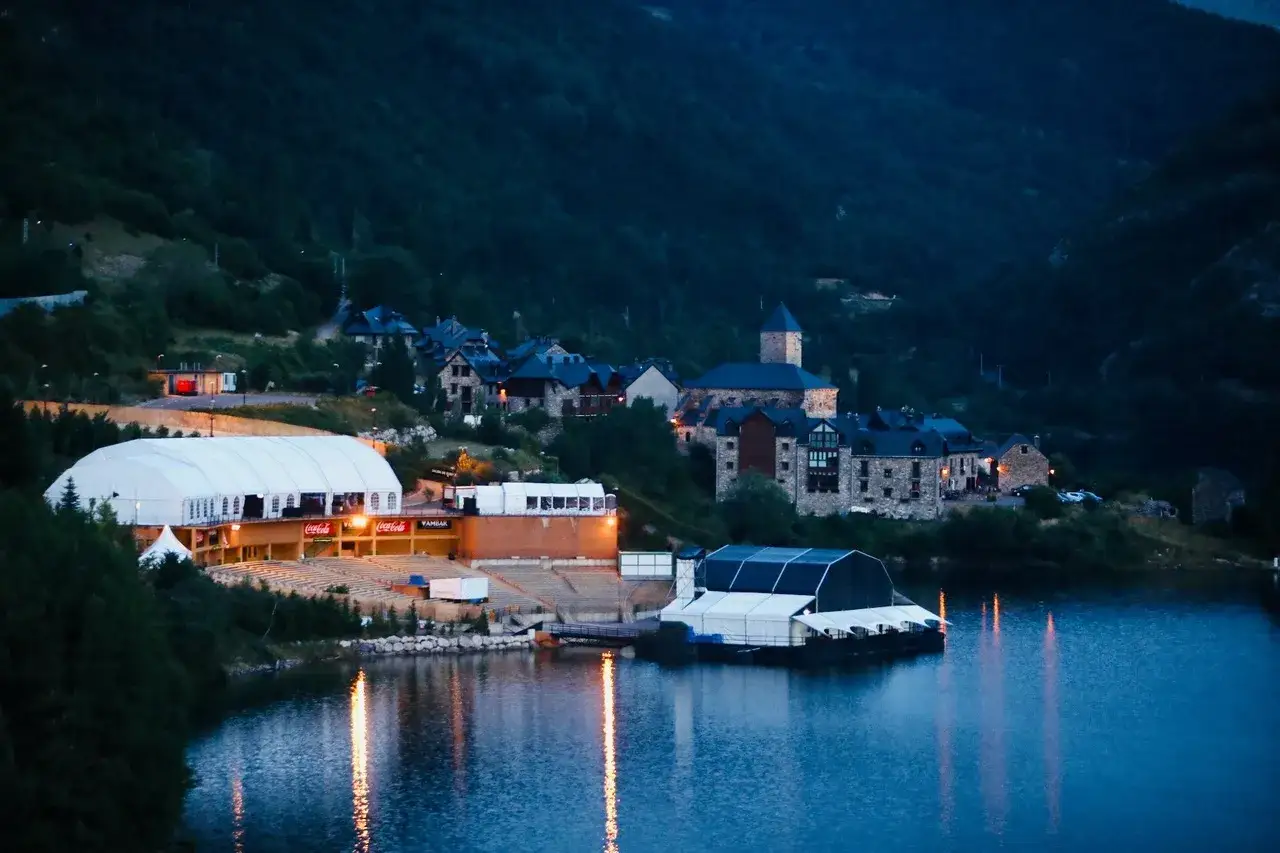 Escenario de carpas y gradas junto a un lago, con un pueblo en la ladera montañosa. Noche de festival en el Pirineo Sur.