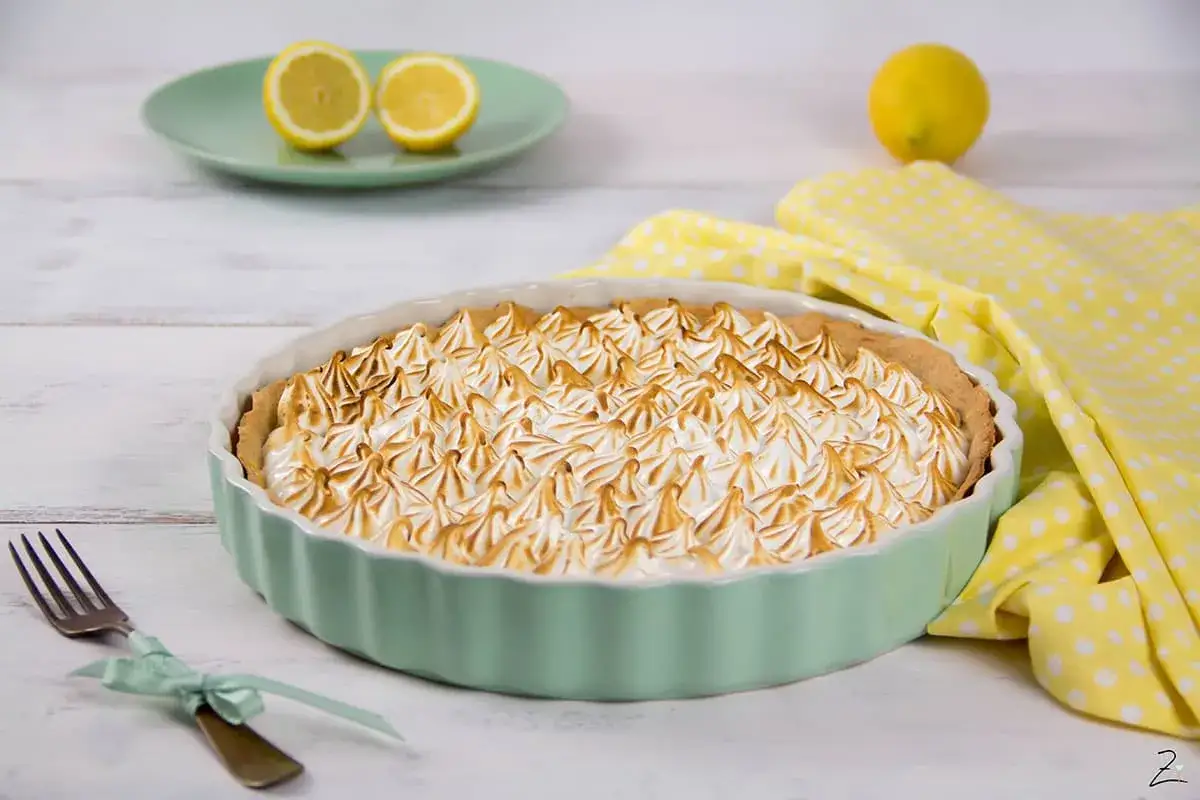 Eine köstliche Tarte au Citron mit gebackener Baiserhaube in einer mintgrünen Form, daneben Zitronen und ein gelbes Tuch.