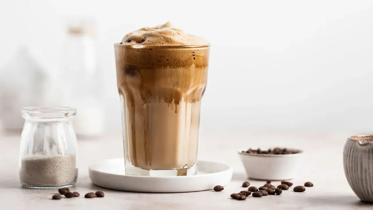 Un bicchiere di crema al caff&egrave; in bottiglia, con schiuma densa e caff&egrave; freddo. Vicino, chicchi di caff&egrave; e una piccola bottiglia di zucchero.