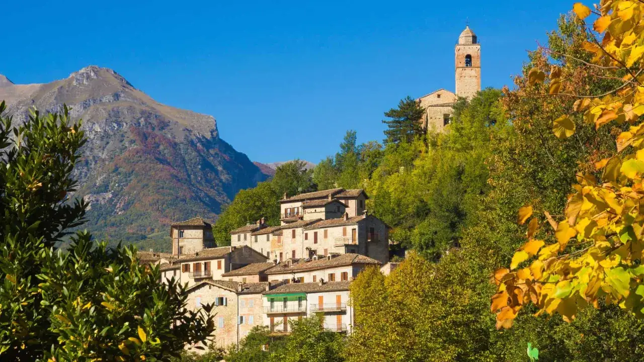 Borgo incantevole nel Val di Norcia, con case in pietra e un campanile che svetta tra gli alberi autunnali dai colori caldi.