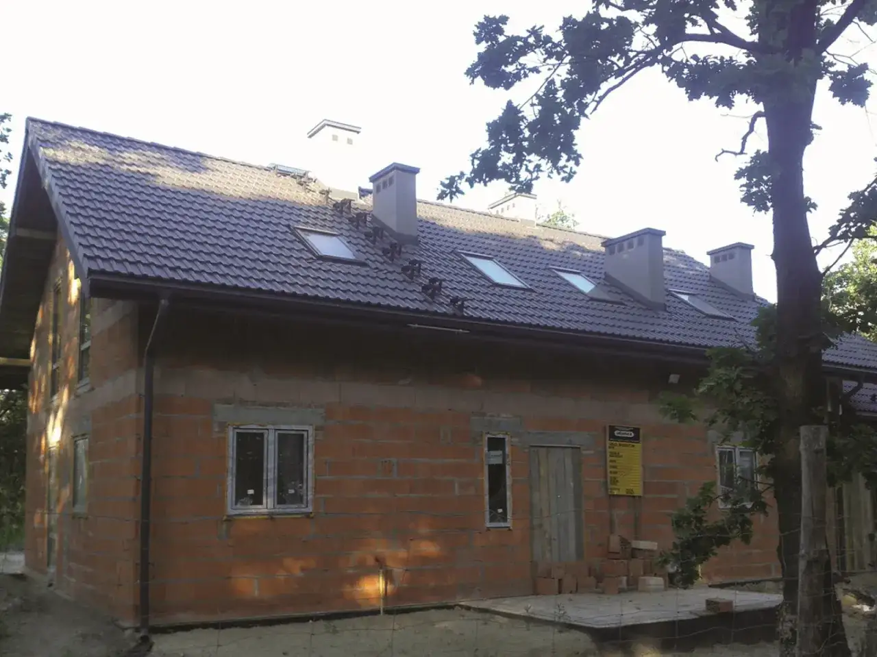 Montaż rury spustowej na nowym domu z cegły. Dach pokryty ciemną dachówką, widoczne okna dachowe i kominy.