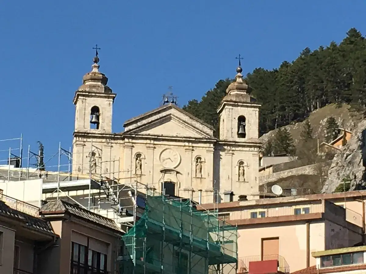 Chiesa di Castel di Sangro, con campanili e orologio, incastonata tra case e montagne boscose.