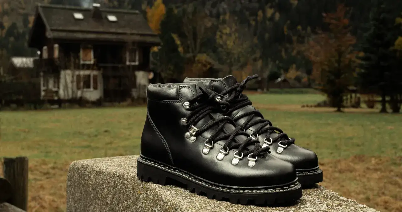 Bottes en cuir noir robustes, parfaites comme chaussure pour marcher dans la neige. Elles sont posées sur une pierre devant une maison de montagne.