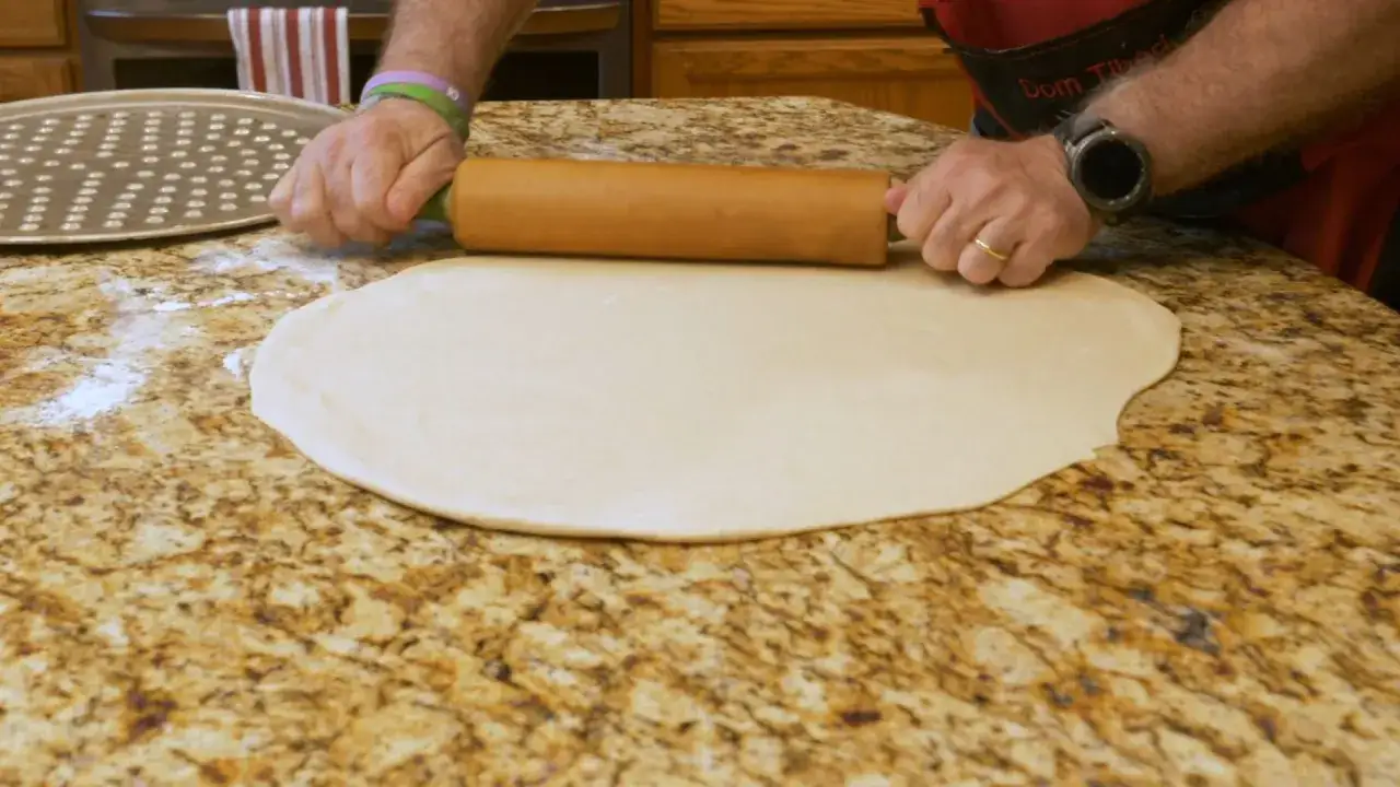 Hände rollen mit einem Nudelholz den **Pizzateig aus** auf einer Marmorarbeitsplatte. Ein Pizzablech liegt daneben.