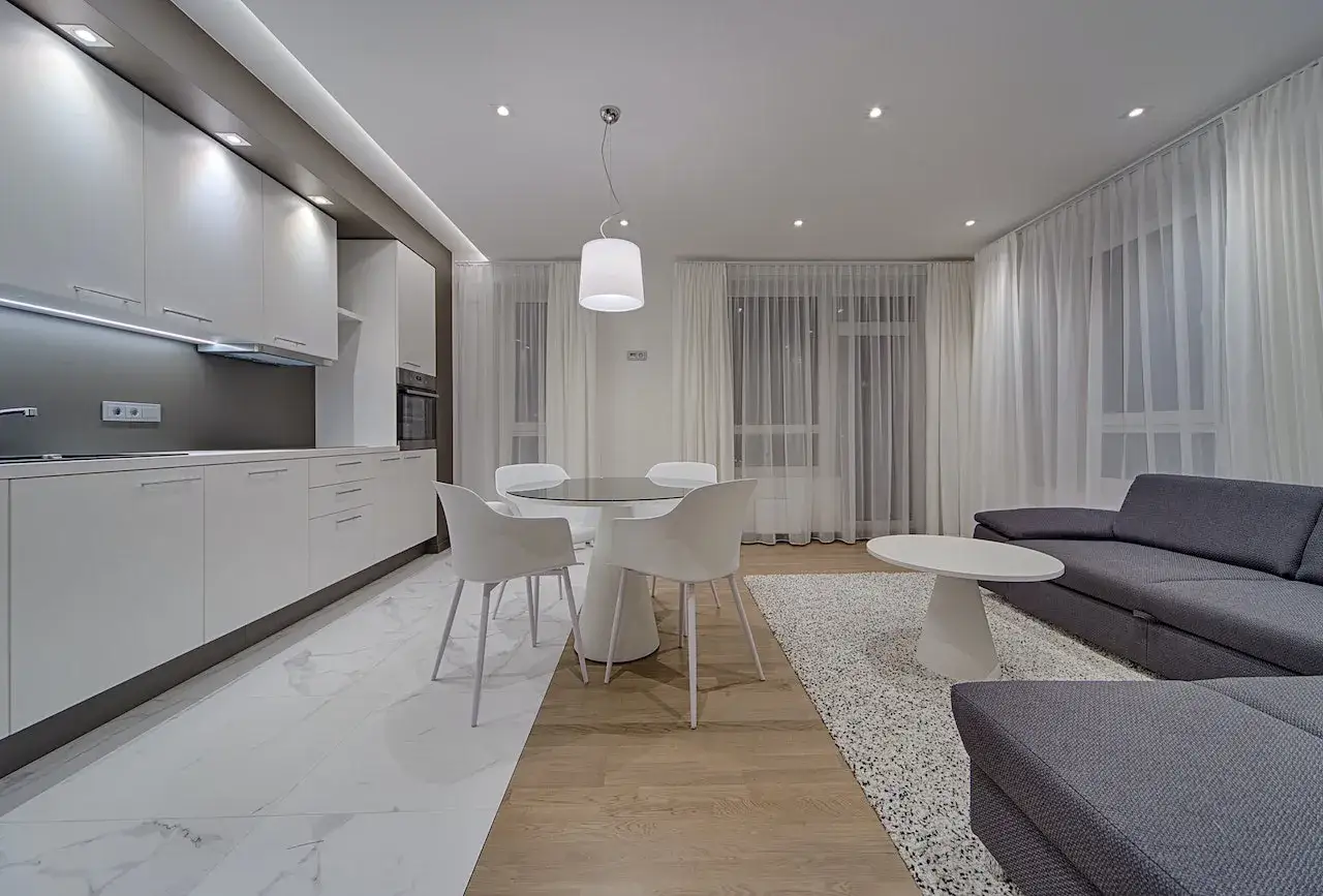 Cuisine et salon modernes aux tons gris anthracite et beige. Canapé d'angle, table ronde, tapis blanc et cuisine épurée.