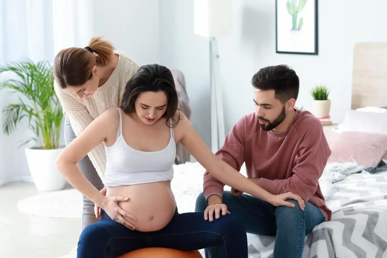 Kobieta w zaawansowanej ciąży, siedząc na piłce, czuje skurcze. Partner i doula wspierają ją, gdy brzuch opada przed porodem.