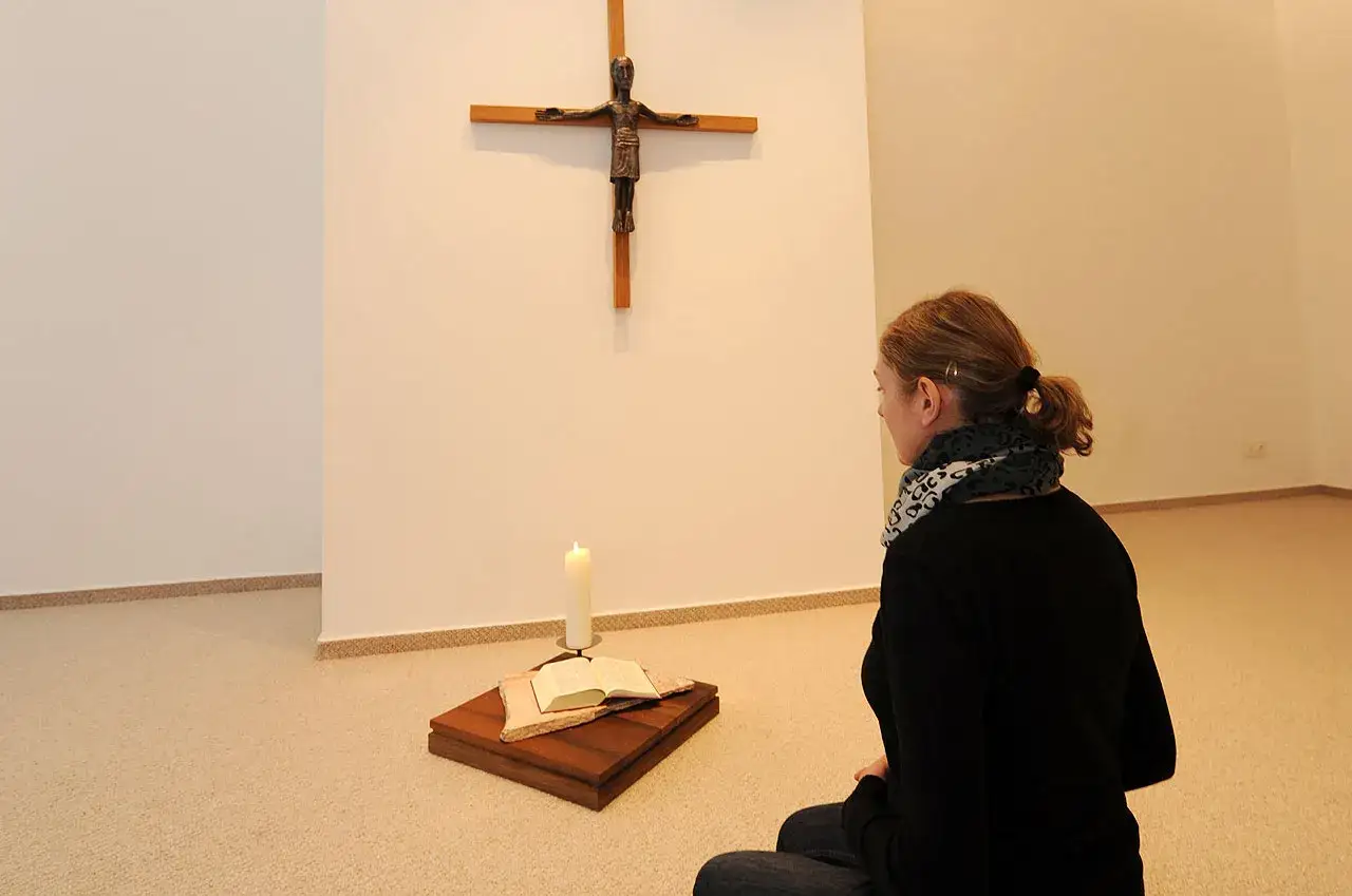 Frau kniet vor einem Kreuz, Kerze und Bibel. Sie sucht Trost und m&ouml;chte an Gott glauben.