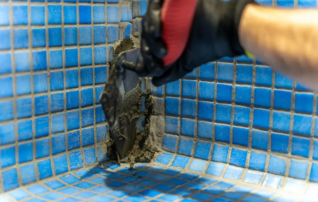 Aplicando mortero para impermeabilizar piscina. Un guante negro con mango rojo usa una espátula para extender el material sobre las baldosas azules.