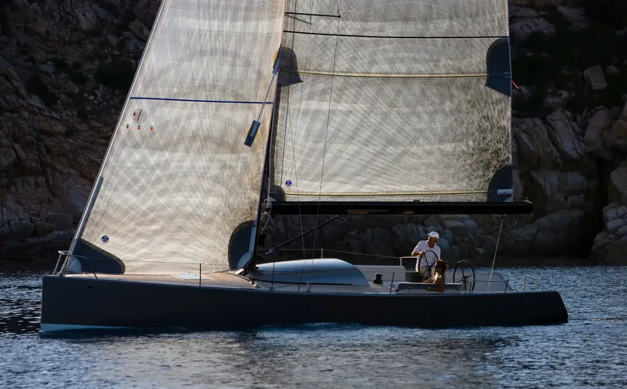 Un elegante **yacht Brenta** scivola sull'acqua blu, con le vele trasparenti illuminate dal sole. Due persone a bordo godono della navigazione.