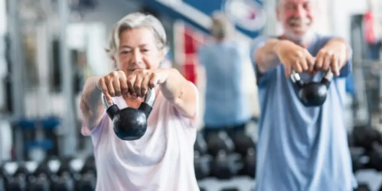 Un couple de retraités souriants participe activement à un programme de mise en forme 60 ans et plus, soulevant des kettlebells avec détermination.