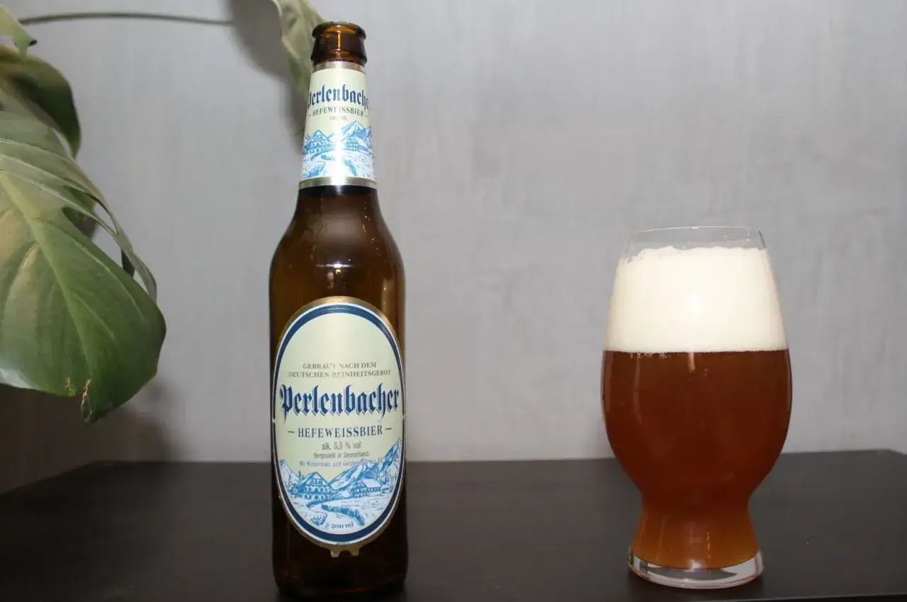 Perlenbacher Hefeweissbier, złociste piwo pszeniczne z obfitą pianką, obok butelki i zielonego liścia.