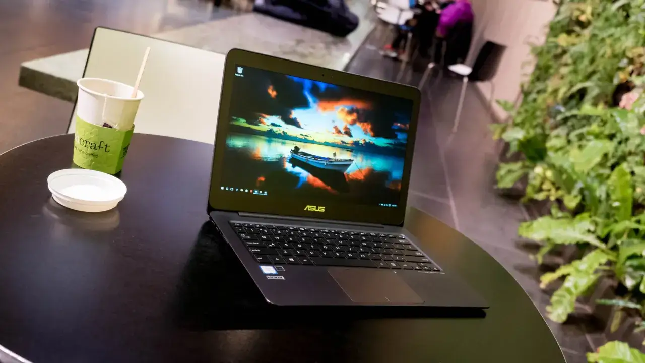 En Asus Zenbook UX305 står på ett mörkt bord bredvid en kopp med te och en grön växtvägg.