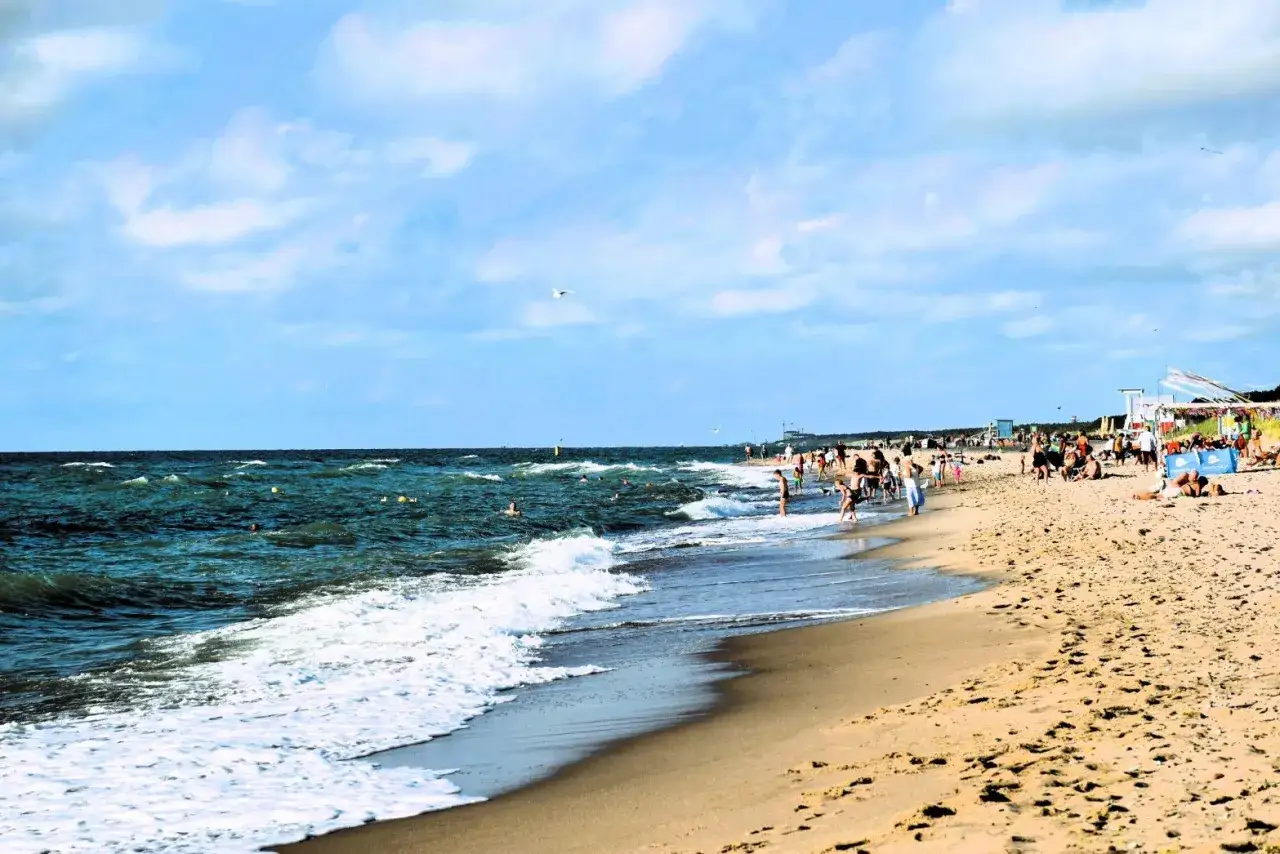 Słoneczny dzień na plaży w Dąbkach. Ludzie kąpią się w morzu i odpoczywają na piasku. Widać budki plażowe i mewy na niebie.