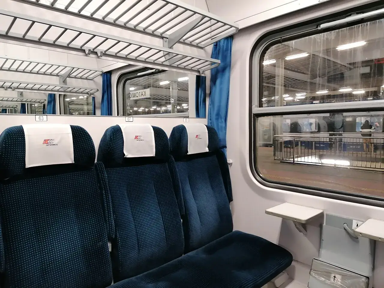Wnętrze wagonu PKP Intercity TLK. Siedzenia w kolorze granatowym, p&oacute;łki nad nimi. Za oknem peron.