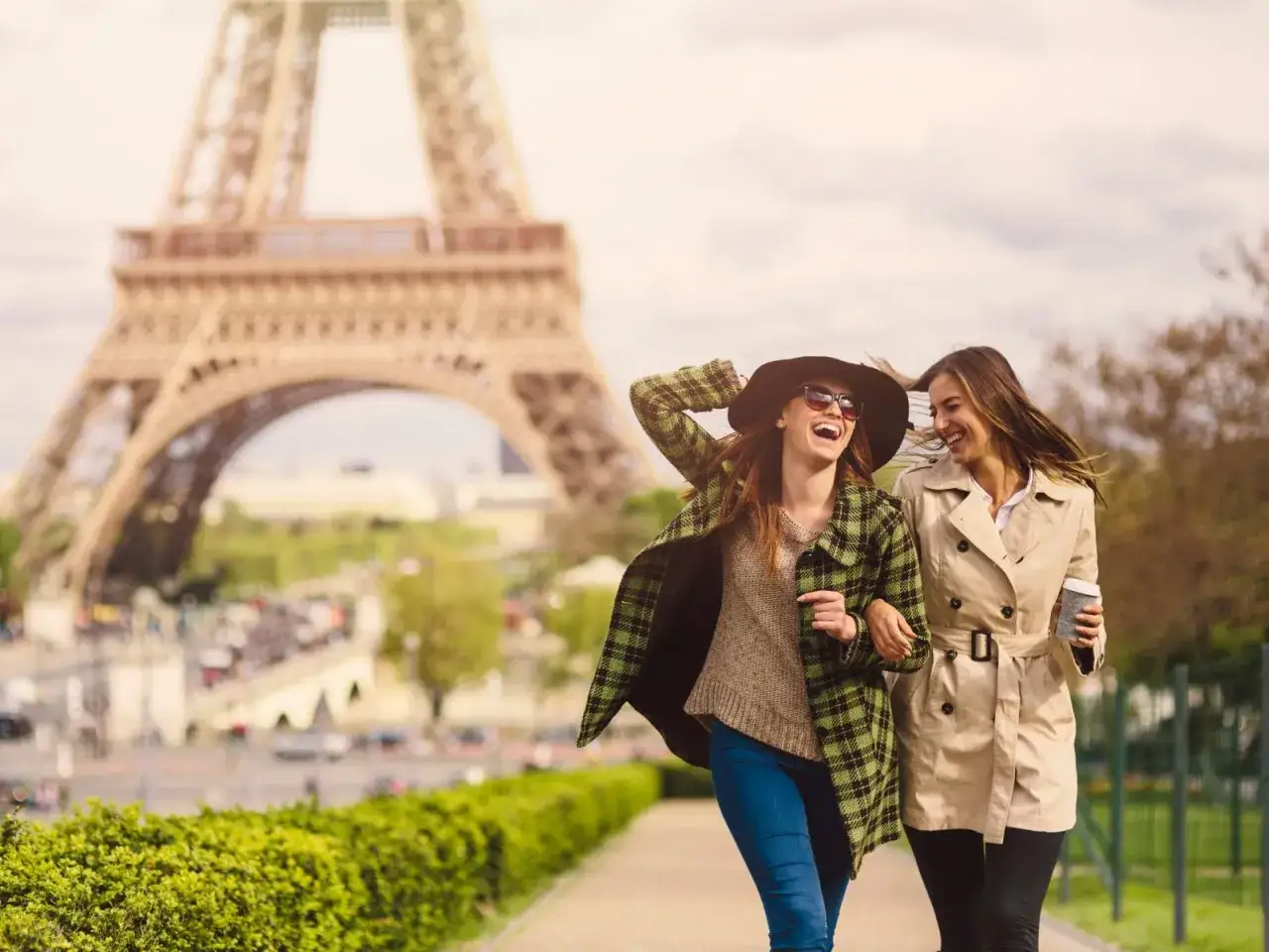 Due amiche ridono passeggiando a Parigi, con la Torre Eiffel sullo sfondo. Perfetto per capire cosa mettere in valigia per Parigi: cappotti leggeri e un cappello!