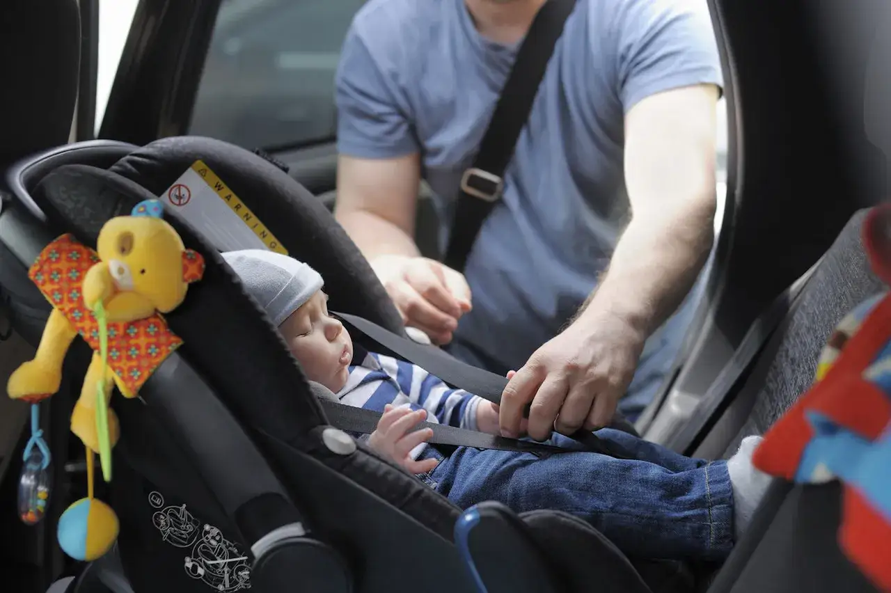 Un padre ajusta el arnés de seguridad de su bebé en la silla del coche. El bebé duerme plácidamente, listo para viajar. Es importante saber cuándo puede ir un niño delante, pero este pequeño va seguro atrás.