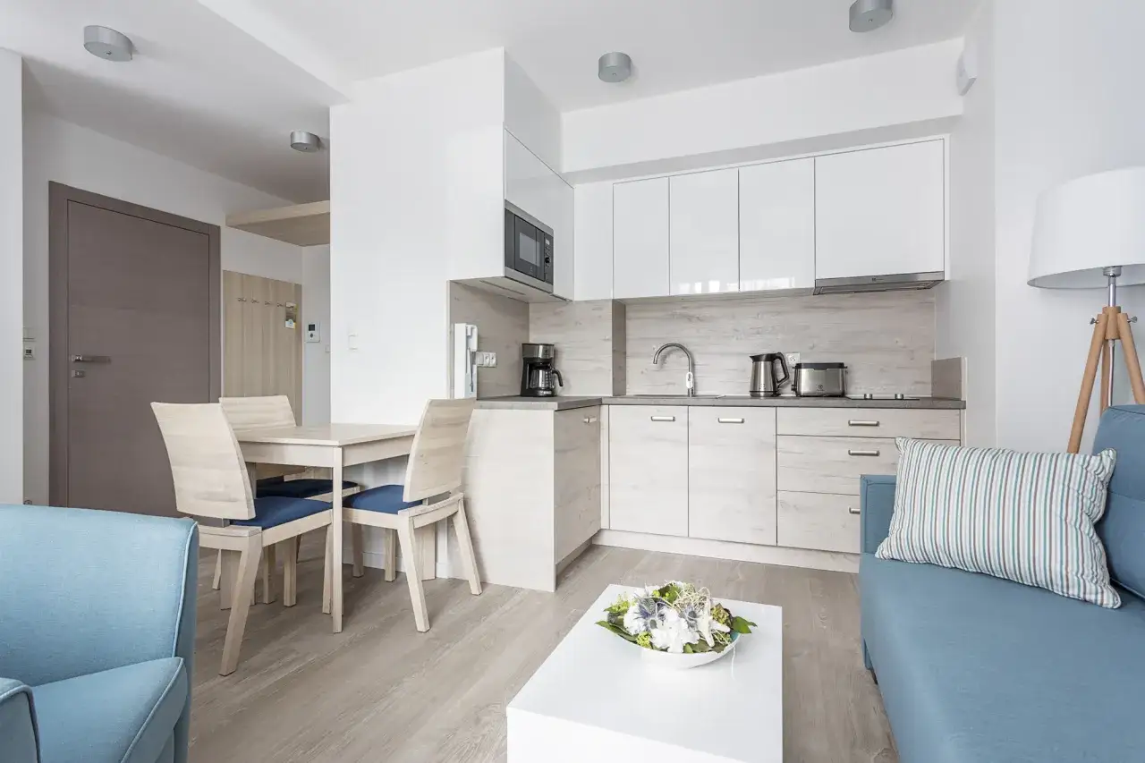 Nowoczesne apartamenty przy morzu. Jasna kuchnia z białymi frontami, jadalnia z drewnianymi krzesłami i salon z błękitną kanapą.