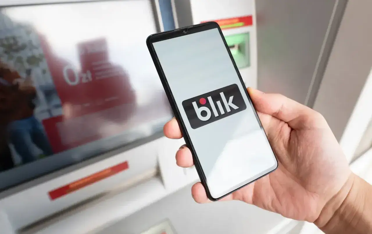 Dłoń trzyma smartfon z logo Blik przed bankomatem. Kto wymyślił Blik? To polski system płatności.