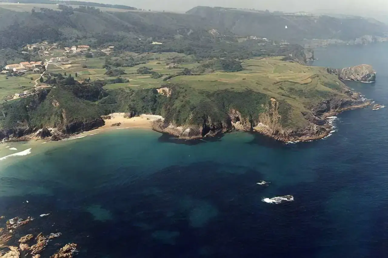 Vista a&eacute;rea de la **playa de Aramal**, con acantilados verdes, arena dorada y aguas turquesas. Un pueblo se asoma entre las colinas.