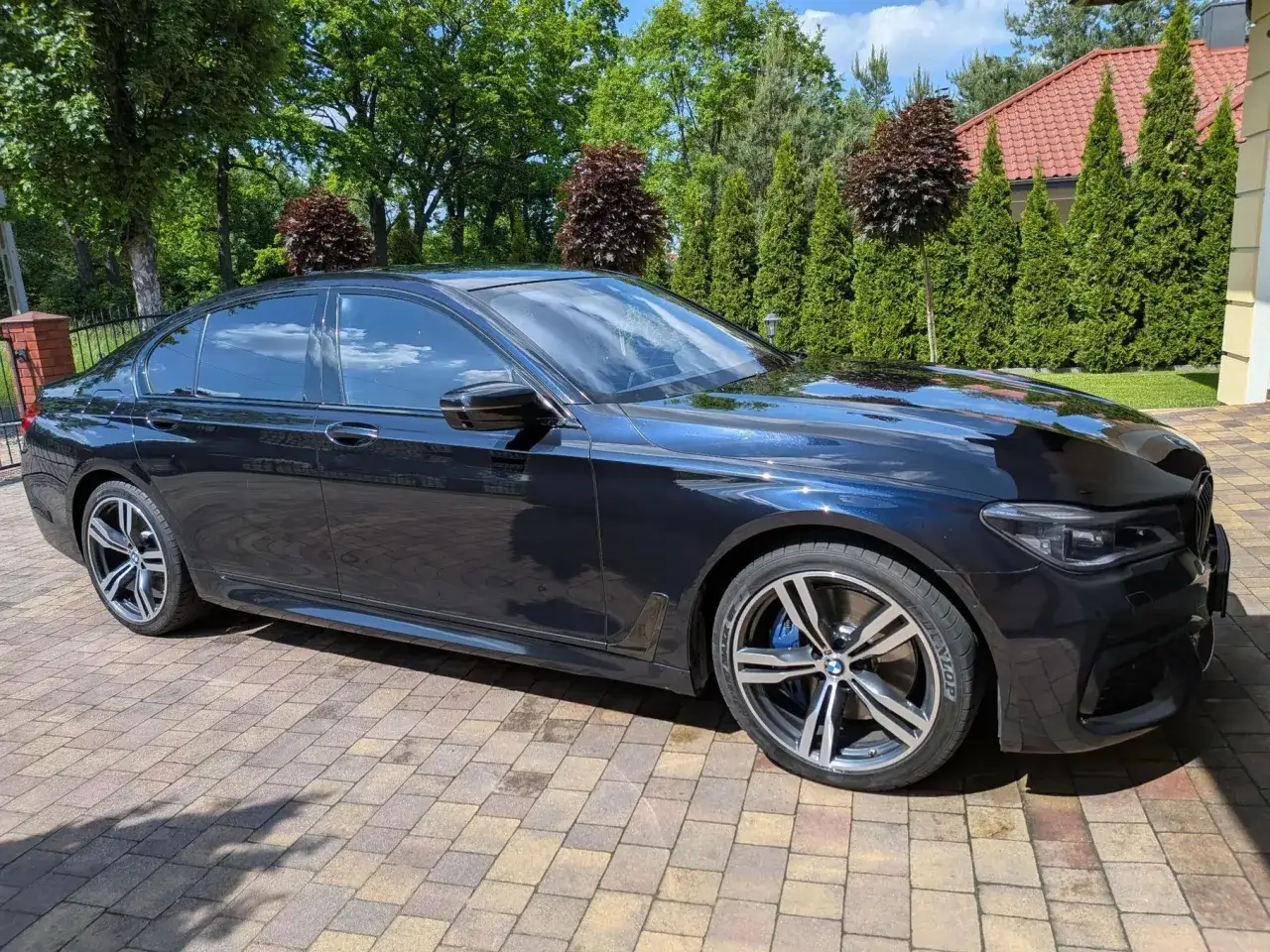 Lśniące, ciemne bmw 730d zaparkowane na brukowanym podjeździe, otoczone zielenią i drzewami.