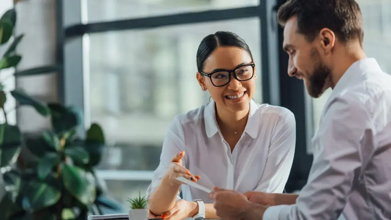 A woman with glasses smiles while discussing a document, possibly related to her PCC certification, with a man in a bright office setting.