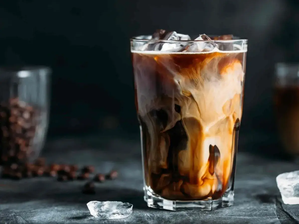 Un bicchiere di latte macchiato freddo con striature di latte e caffè, cubetti di ghiaccio e chicchi di caffè sullo sfondo.