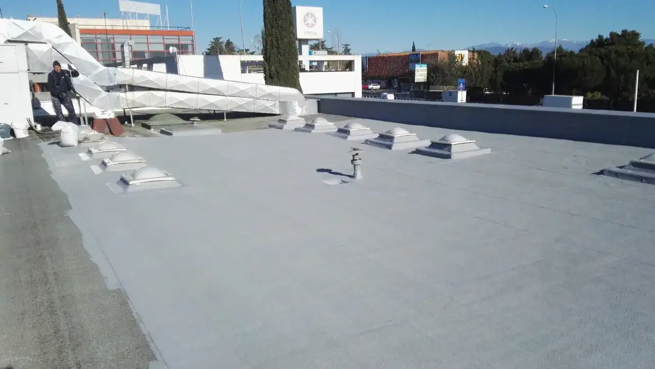 Trabajador aplicando pintura para impermeabilizaci&oacute;n de tejados. Se ven claraboyas y conductos de ventilaci&oacute;n en la azotea.
