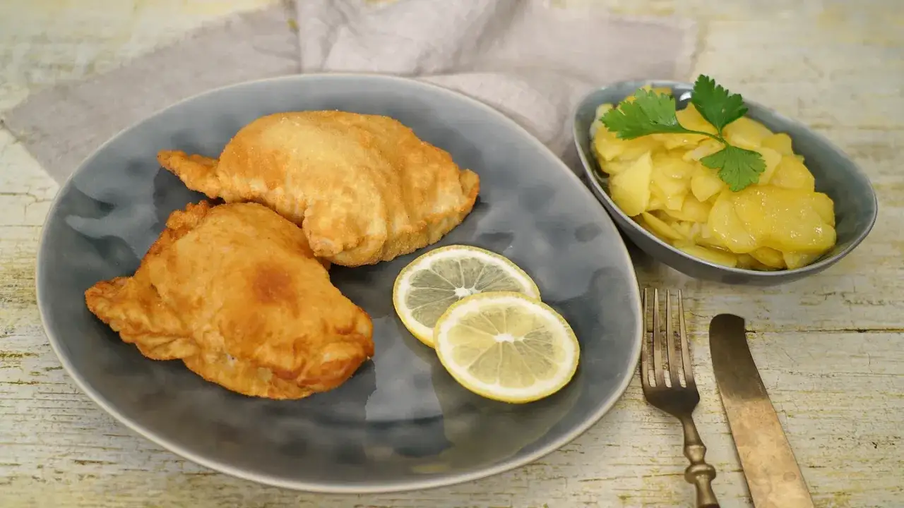 Zwei goldbraune Wiener Schnitzel auf einem grauen Teller mit Zitronenscheiben und Kartoffelsalat daneben.