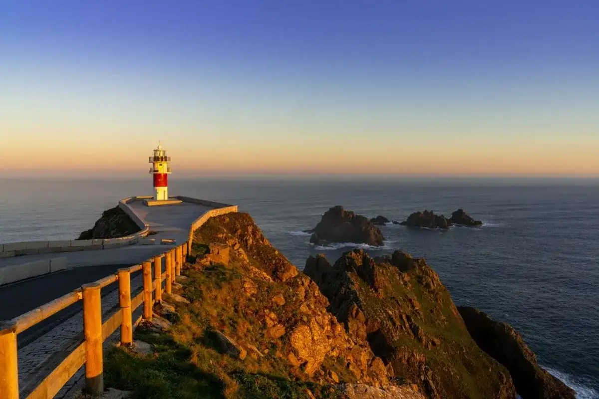 El faro de estaca de bares se alza sobre un acantilado rocoso al atardecer, vigilando el mar azul y las islas cercanas.