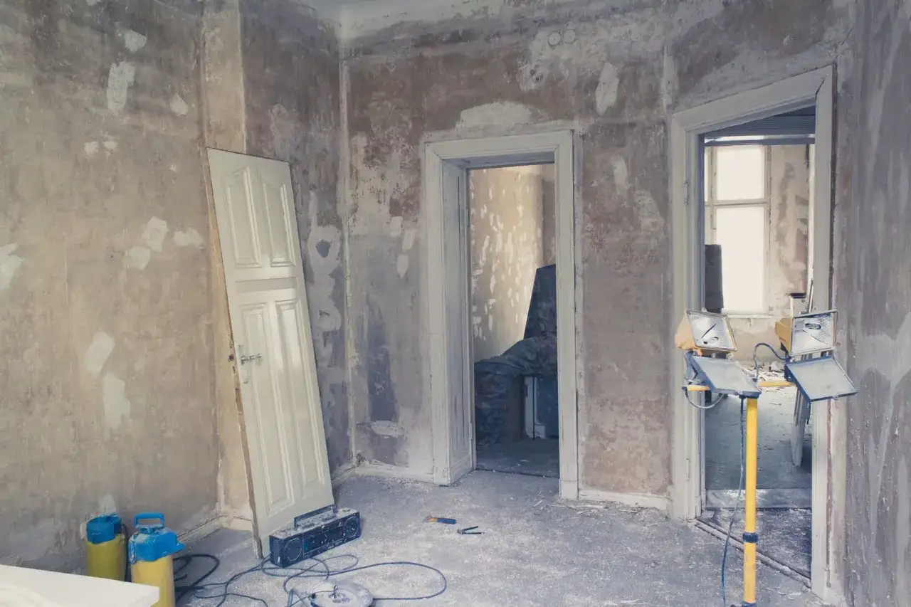 Salle en rénovation, murs bruts prêts à recevoir un enduit sur peinture. Une porte blanche appuyée, des outils et des projecteurs.