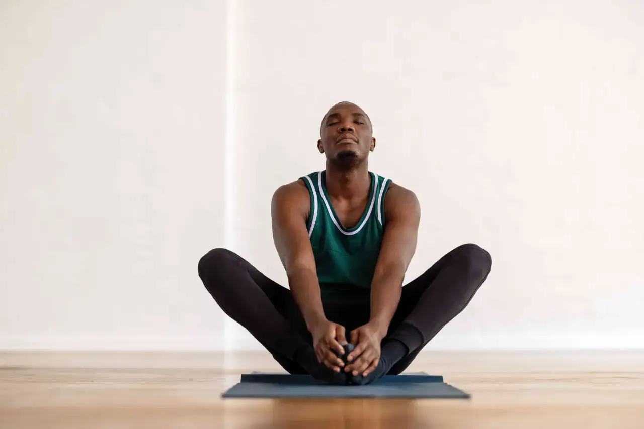 Man i grön linne och svarta träningskläder sitter på en yogamatta i fjärilsposition, med slutna ögon. En avslappnande ställning som kan hjälpa till att få ut gaser ur magen snabbt.