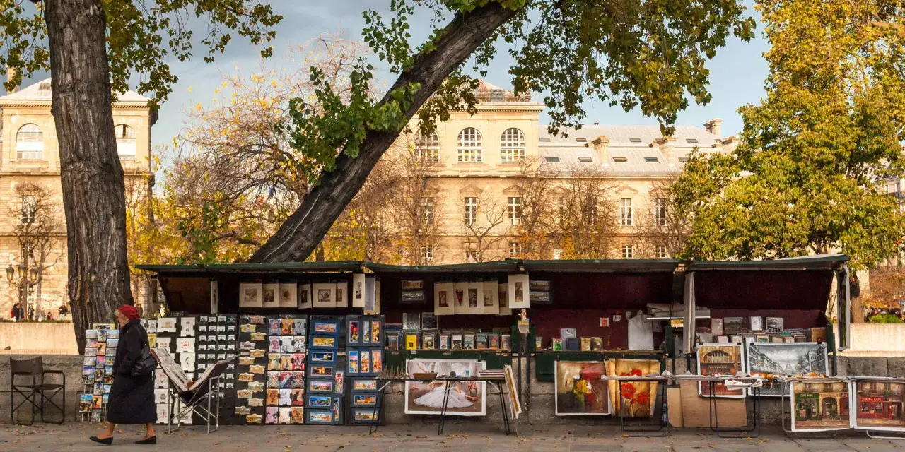 Ein **Flohmarkt in Paris** mit Büchern, Postkarten und Gemälden. Eine Frau im roten Kopftuch schlendert vorbei.