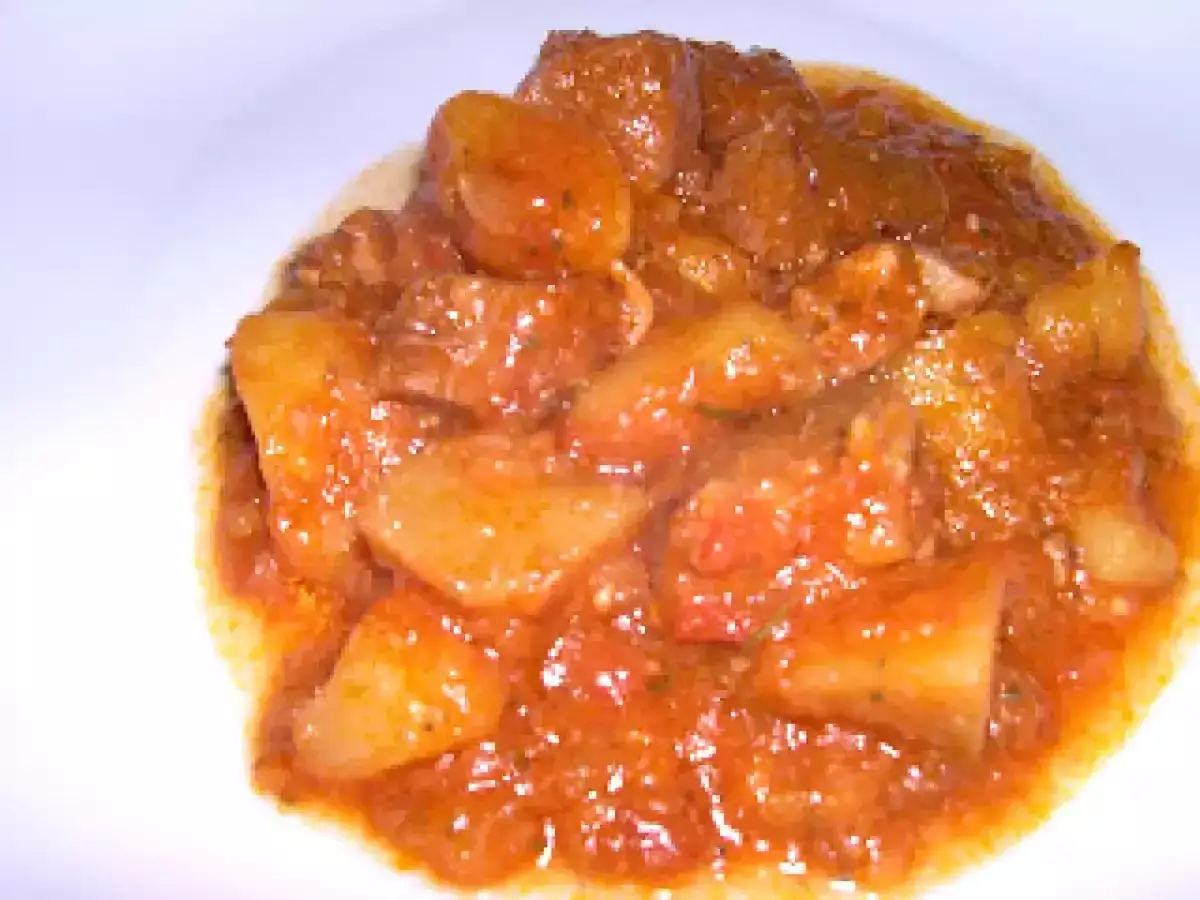 Un saporito spezzatino di maiale con patate, cotto lentamente in un sugo rosso e aromatico, servito in un piatto bianco.