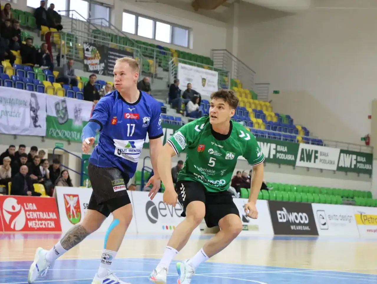 Wiktor Kwiatkowski w akcji na boisku. Zawodnik w niebieskiej koszulce z numerem 17 i zielonej z numerem 5 walczą o piłkę.