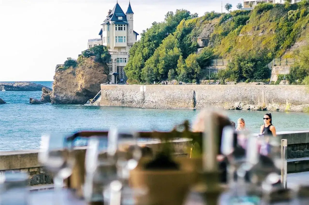 Vue sur un ch&acirc;teau sur une falaise &agrave; Biarritz, avec la mer bleue et des verres en premier plan. Id&eacute;al pour savoir o&ugrave; manger &agrave; Biarritz.