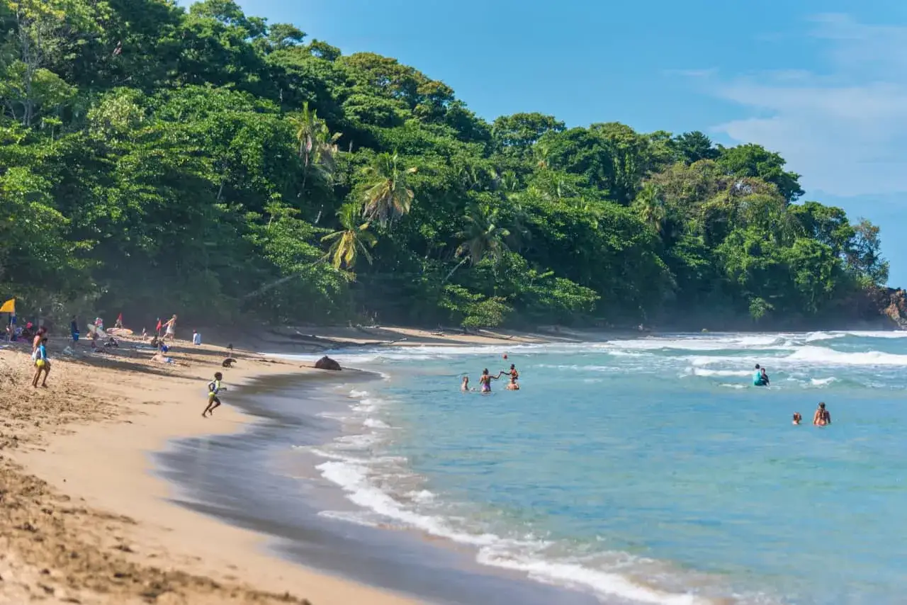 Sonniger Strand in Puerto Viejo mit üppiger Vegetation und Menschen, die im türkisfarbenen Meer planschen.