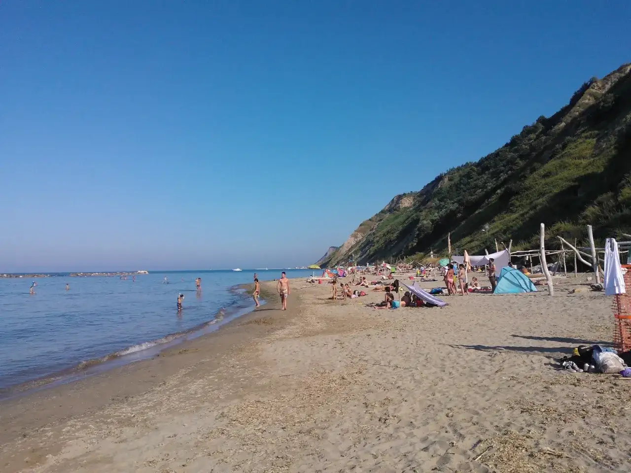 Giornata soleggiata sulla pesaro strand, con bagnanti che si godono il mare e la sabbia sotto la collina verdeggiante.