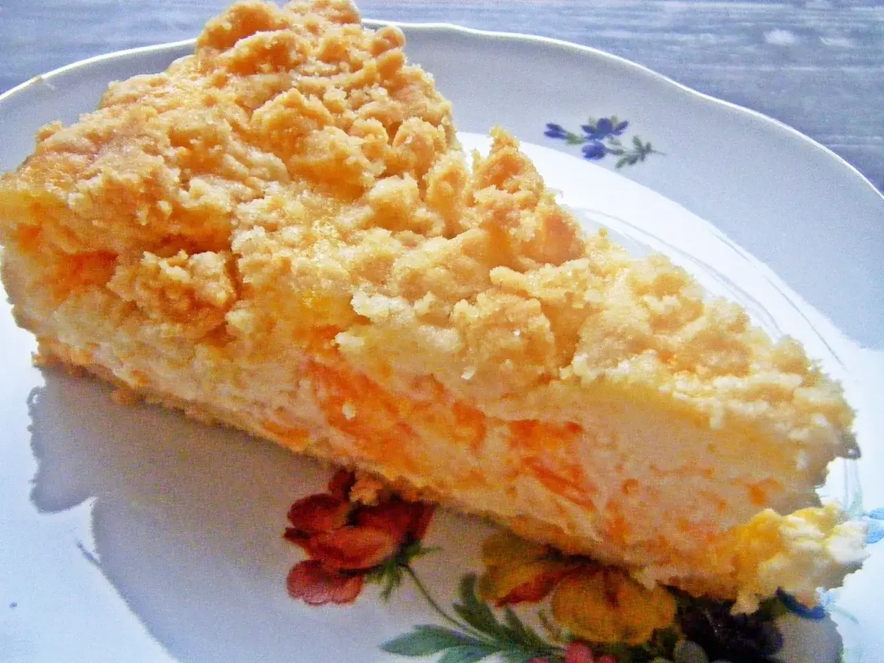 Ein saftiges Stück Mandarinenkuchen vom Blech mit knuspriger Streuseldecke auf einem Teller mit Blumenmuster.