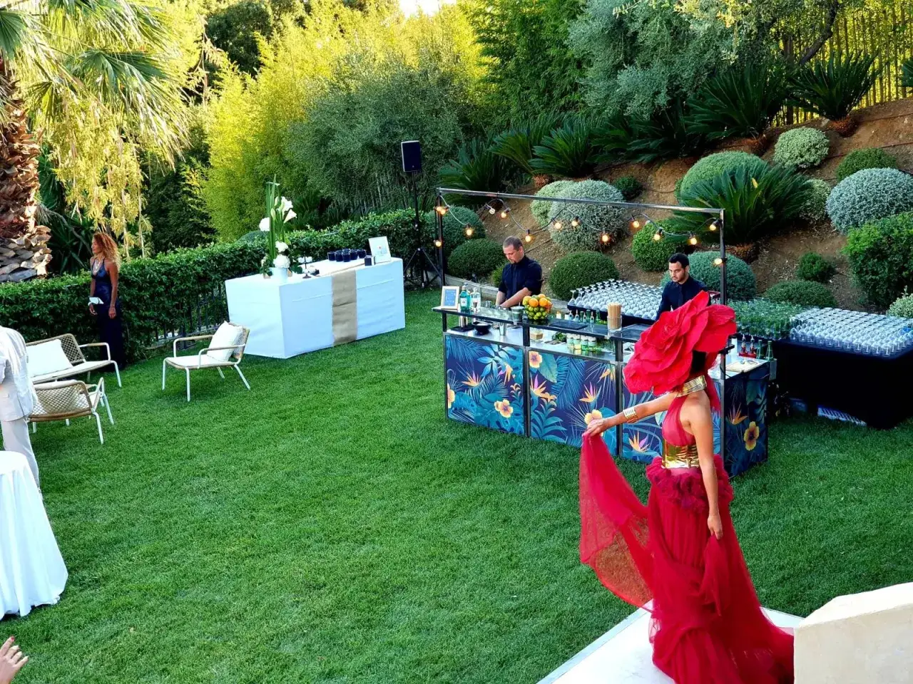 Une femme en robe rouge spectaculaire se tient pr&egrave;s d'un bar tropical, pr&ecirc;te pour un cocktail mariage. Des barmans pr&eacute;parent des boissons dans un jardin luxuriant.