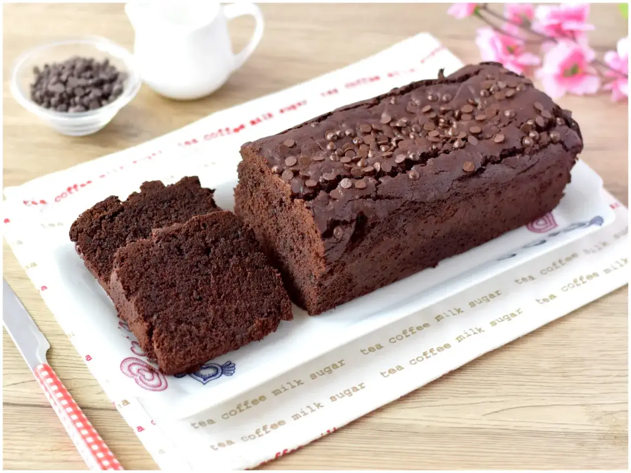 Un goloso plumcake al cioccolato, cosparso di gocce, è affettato su un piatto. Accanto, una ciotolina di gocce di cioccolato e una zuccheriera.