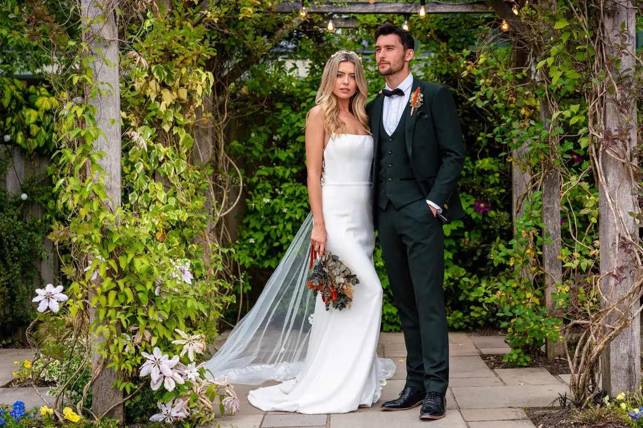 A bride in a white gown stands beside a groom in a dark green suit, a stylish choice that makes him stand out from any potential groomsmen.