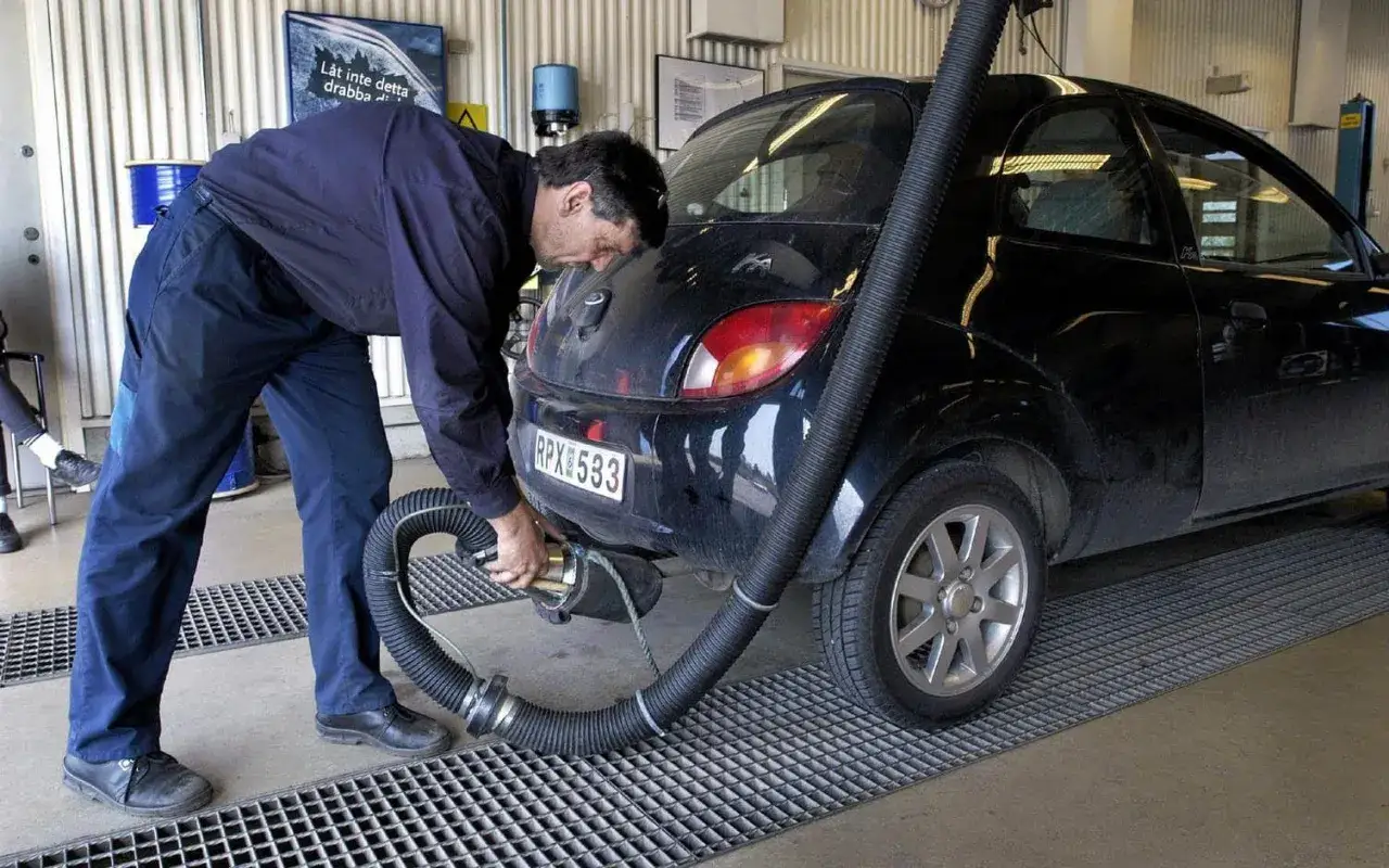 En tekniker ansluter en slang till avgasröret på en svart bil under en besiktning. Detta är en del av kontrollen för att säkerställa att bilen klarar kraven, kanske för att undvika problem med egr delete.