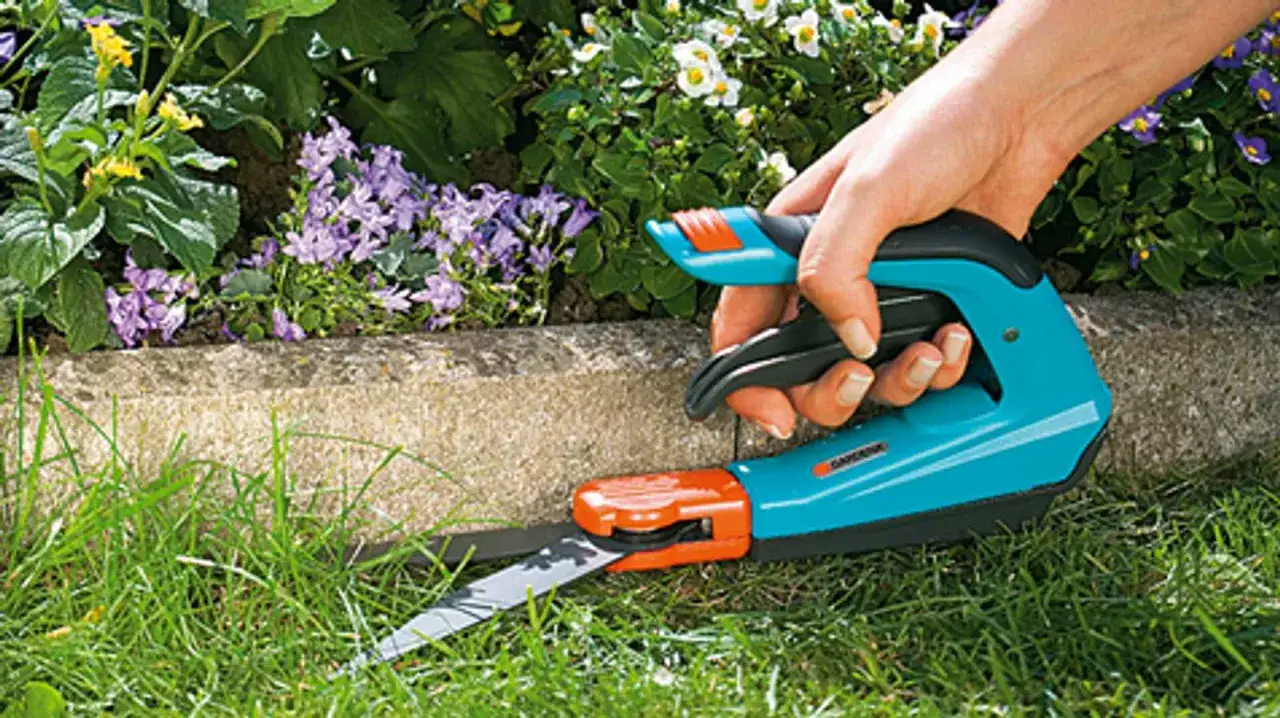 Hand mit Gartenschere schneidet präzise die Rasenkante. Blumen und Grünpflanzen im Hintergrund.