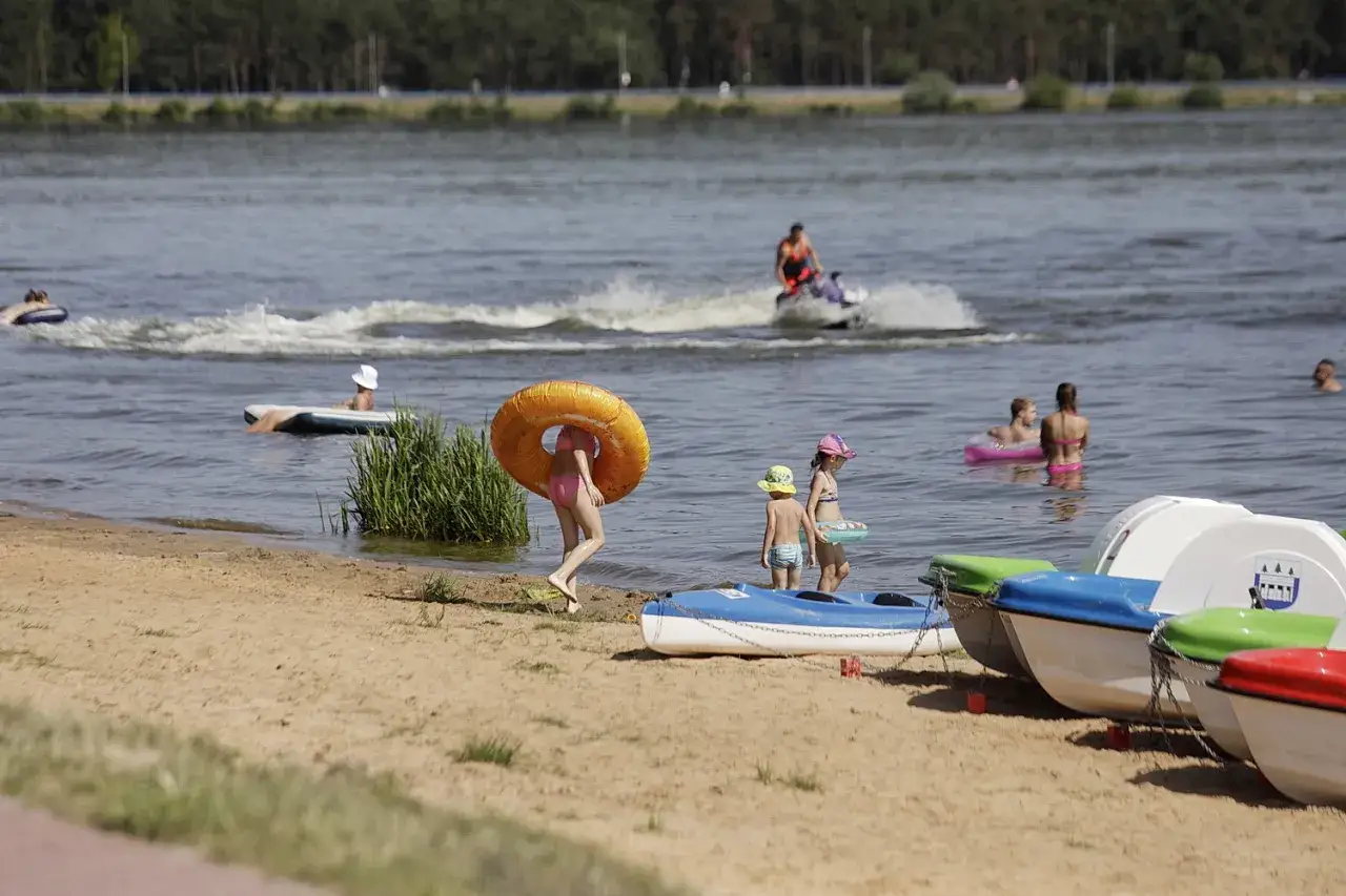 Dzieci bawią się na plaży nad zalewem Brody, jedna dziewczynka niesie koło ratunkowe. W tle widać ludzi pływających i jeżdżących na skuterze wodnym.